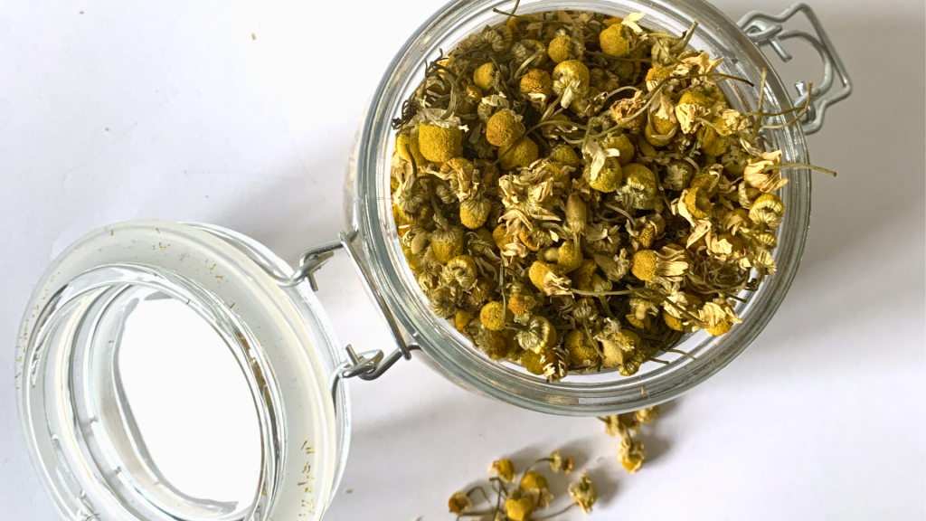 A clear glass jar filled with dried chamomile flowers, showcasing their delicate, golden petals, used for making herbal tea and remedies