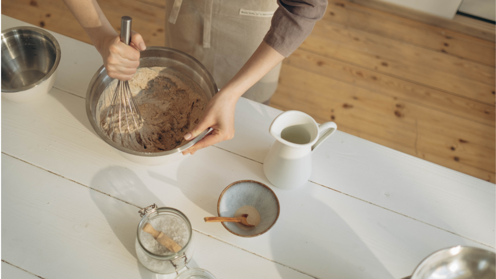 Close-up of hands preparing a vegan cake mix, surrounded by bowls filled with various baking ingredients like flour, sugar, and coconut oil.